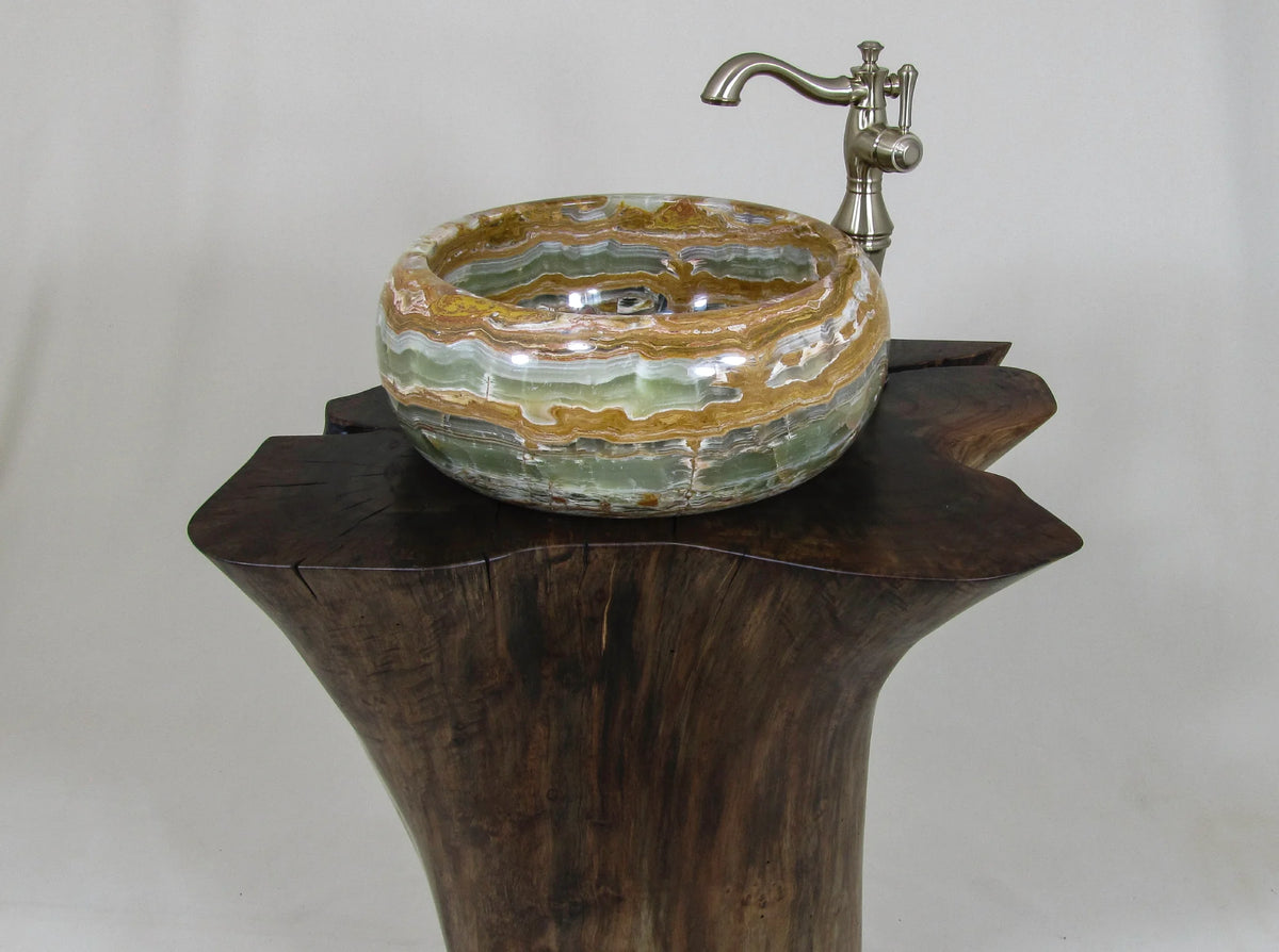 A round onyx vessel sink with bands of brown, gold, green with a silver faucet on a dark wood pedestal.