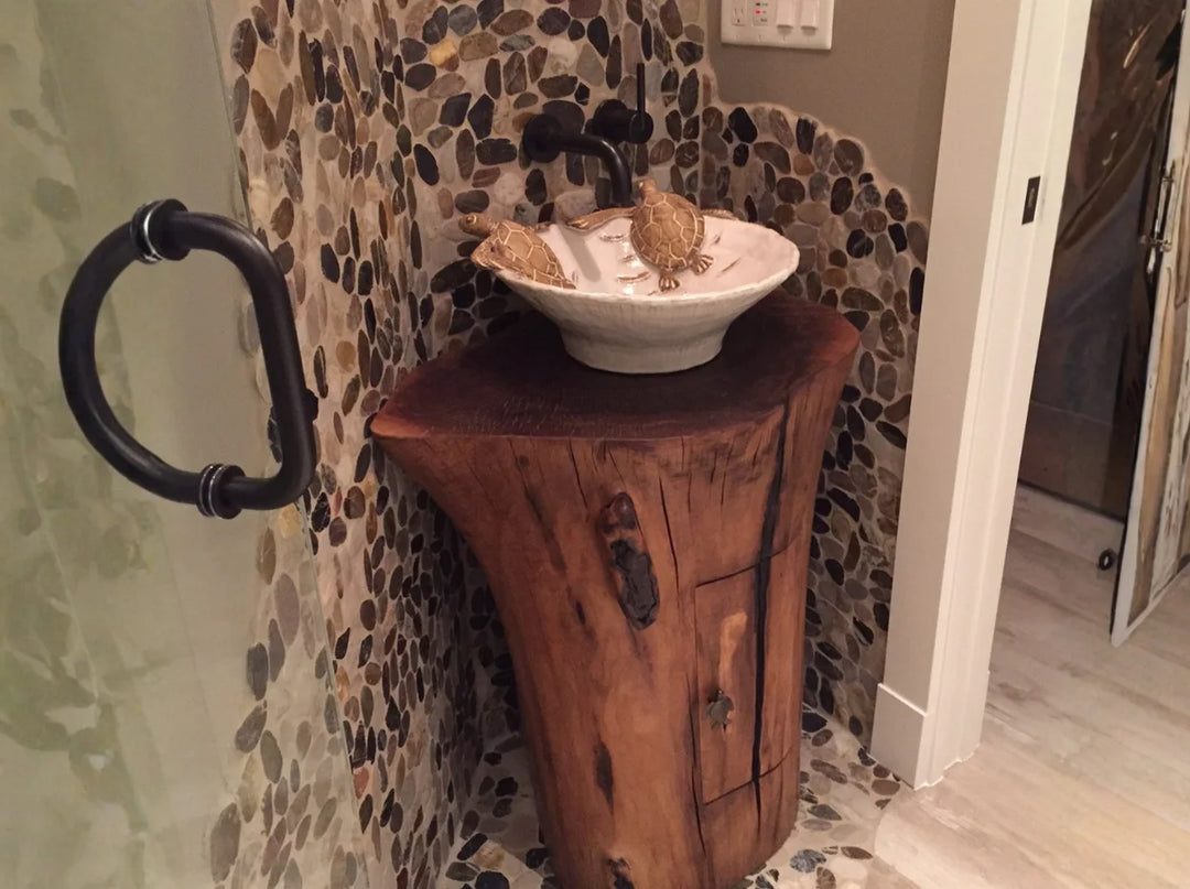 A Black Walnut pedestal sink with a ivory colored vessel with turtles on top.  Back ground is a bathroom with small river stone and glass shower.