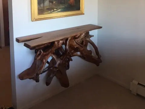 A dark tangled tree root  with a darkened Maple slab on top to serve as a shelf mounted on a white wall  below a picture.