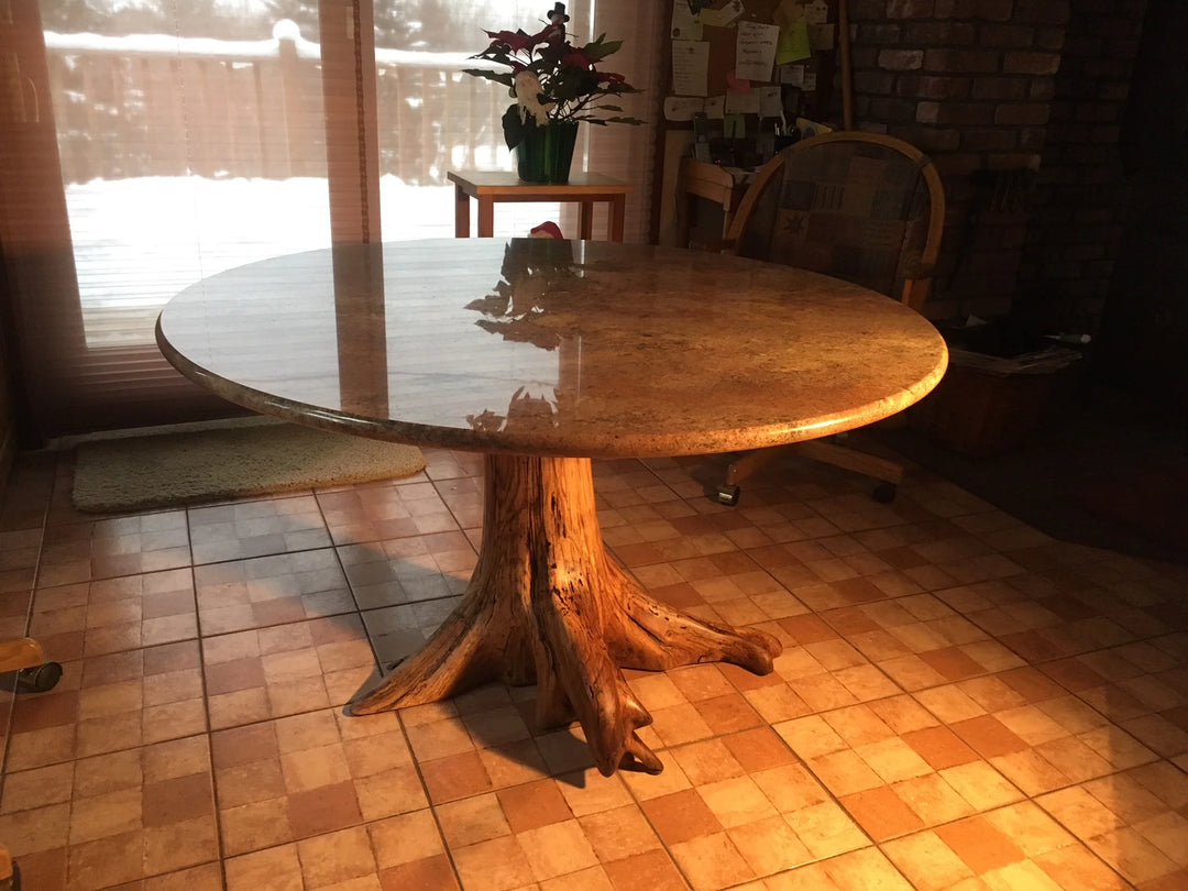 A round granite top dining table with a natural tree-root base, shown in a home dining room with tiled floors and a window in the background.