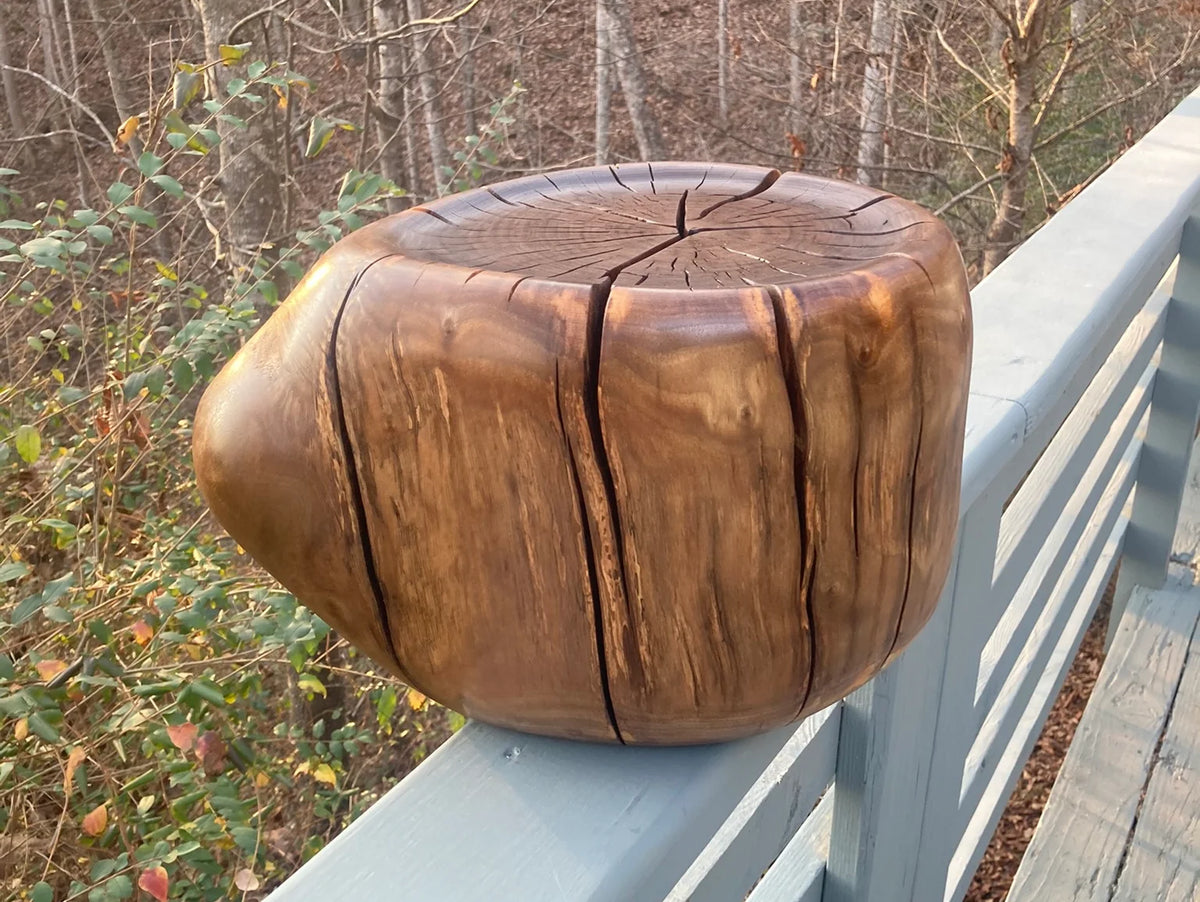 A rustic wooden stump stool with natural grain and a matte finish on a deck rail with a wooded background.