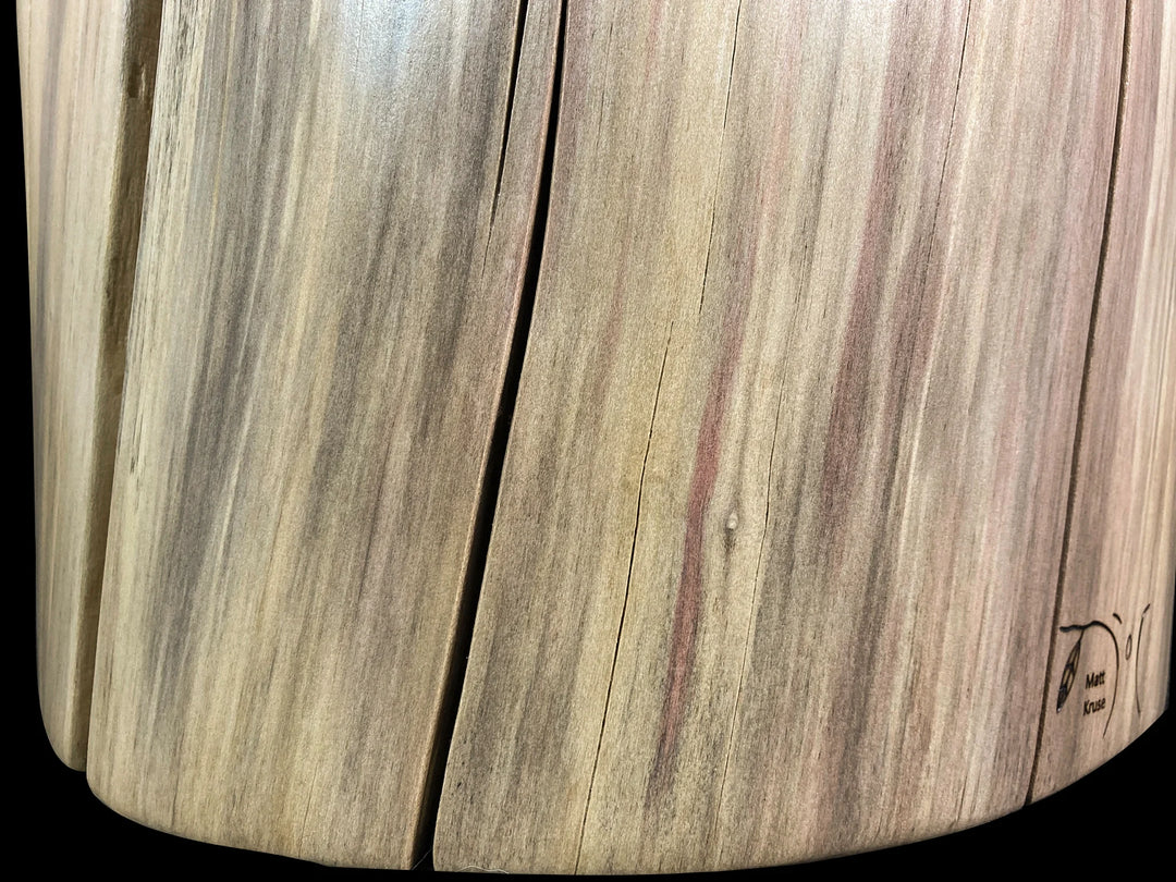 Close-up of a curved wooden surface with visible wood grain, knots, and a small wood brand on the lower right corner, photographed against a black background.