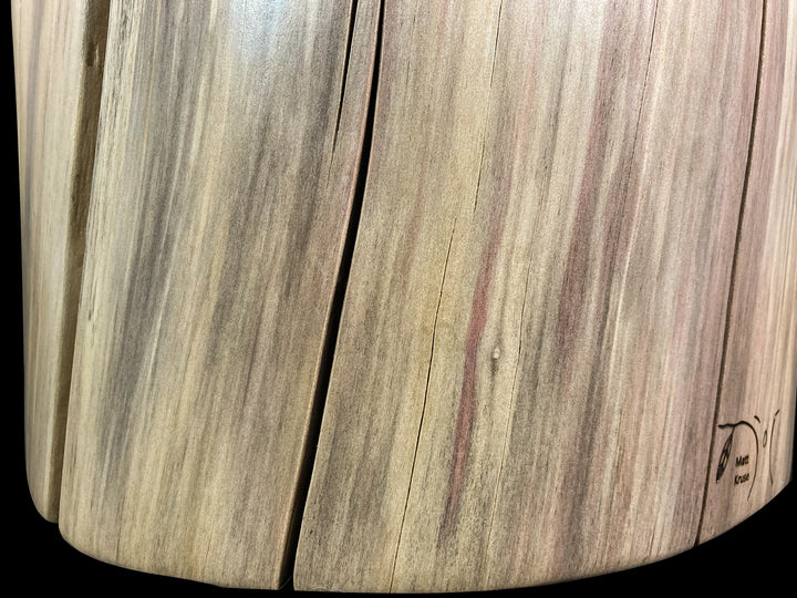 Close-up of a curved wooden surface with visible wood grain, knots, and a small wood brand on the lower right corner, photographed against a black background.
