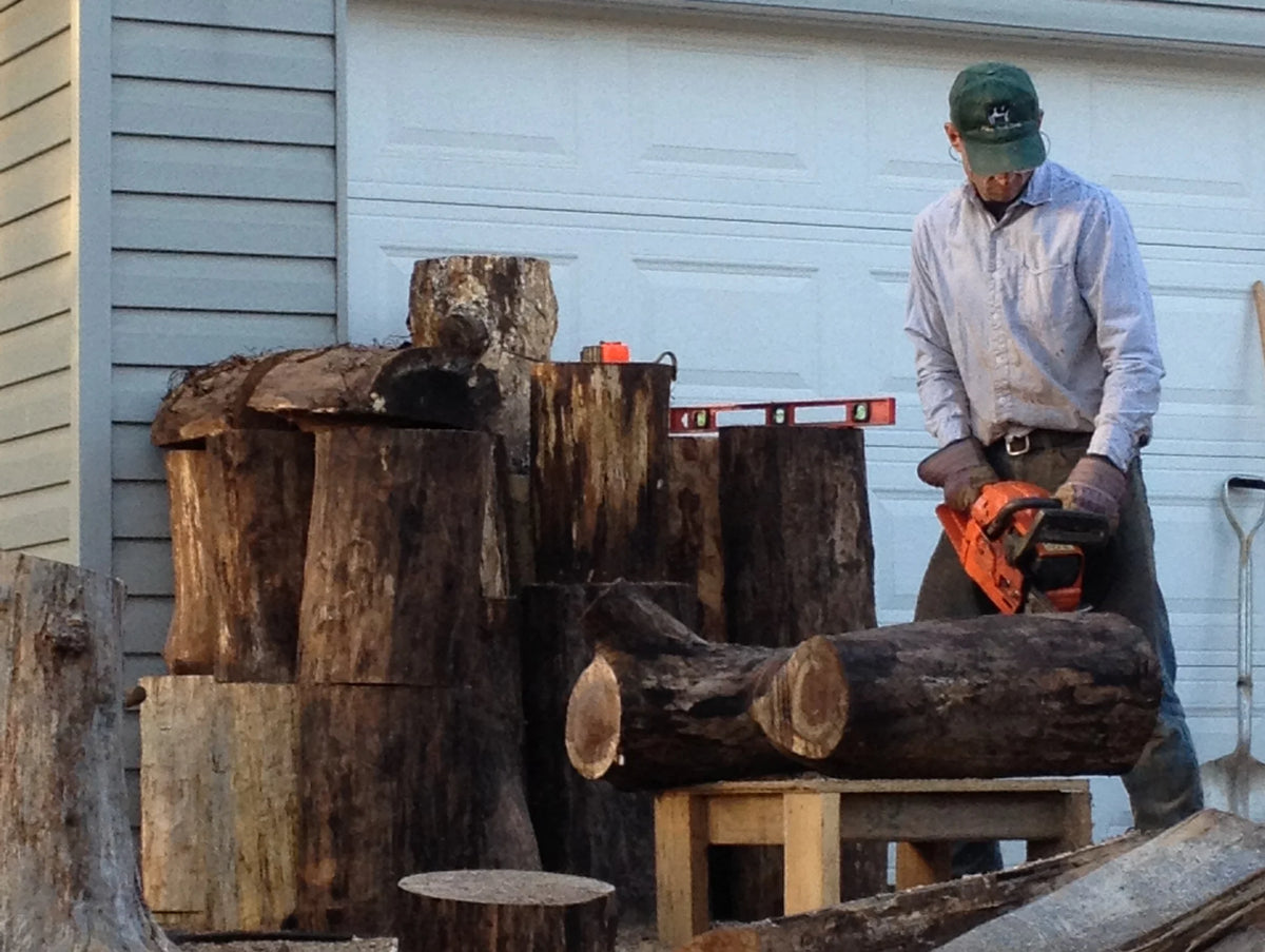 A man wearing a green cap, light blue shirt, and work gloves next to a stack of cut logs cutting with a chainsaw.