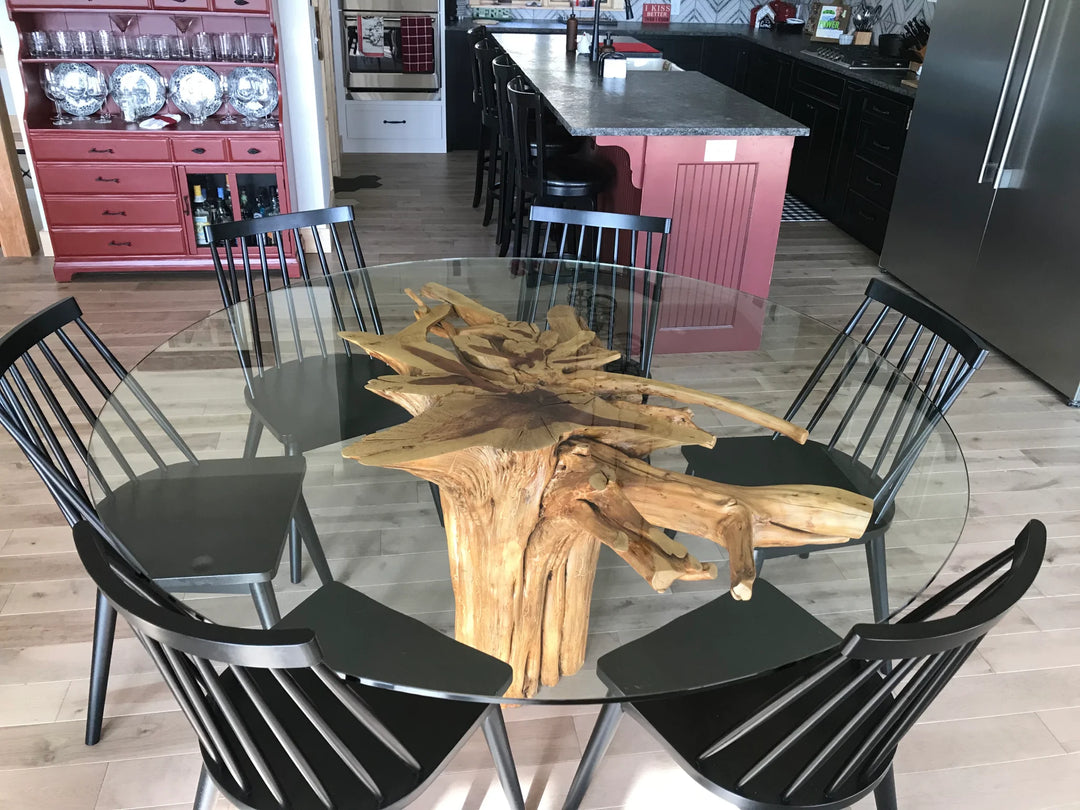 A round glass-top dining table with a large natural tree-root centerpiece, surrounded by six black spindle-back chairs in a showroom setting.