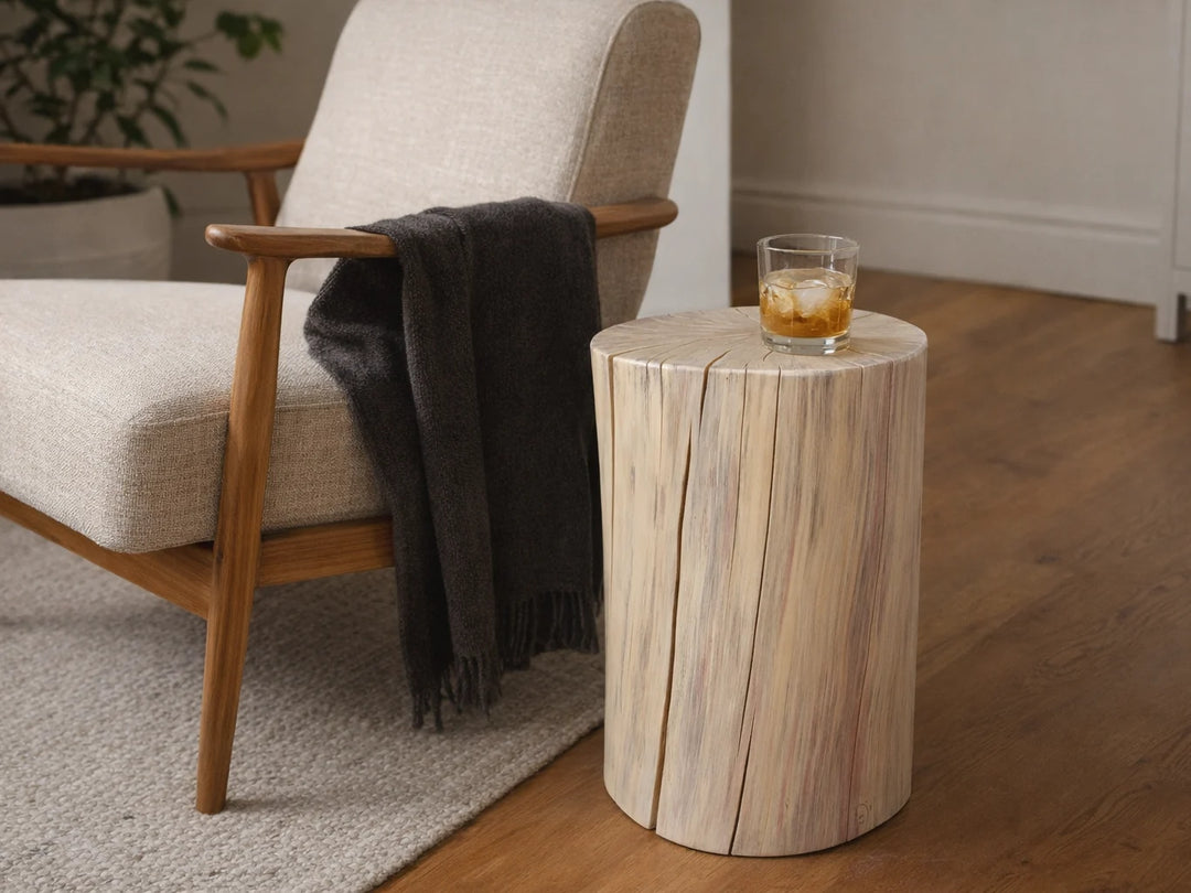A cylindrical side table made from a natural wood stump with visible grain, styled next to a mid-century modern armchair with a dark gray throw blanket and a small glass of iced drink on top.