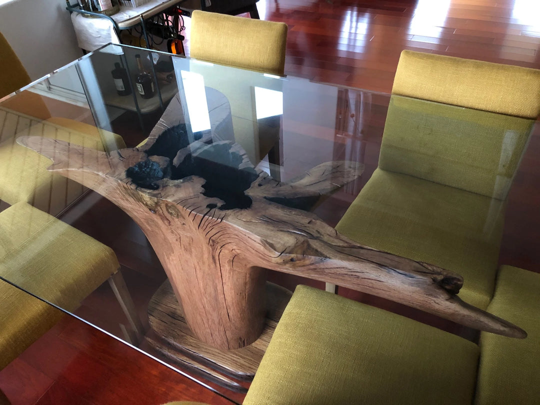 A modern rectangular glass-top dining table supported by a large natural wood root base, surrounded by olive green upholstered chairs in a home setting with a polished wood floor.