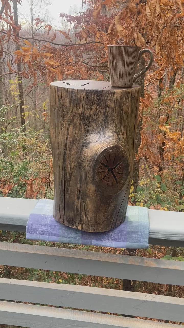 Video of an oak log side table with a large branch knot, sitting on a grey deck rail with a fall time forest in the back ground.